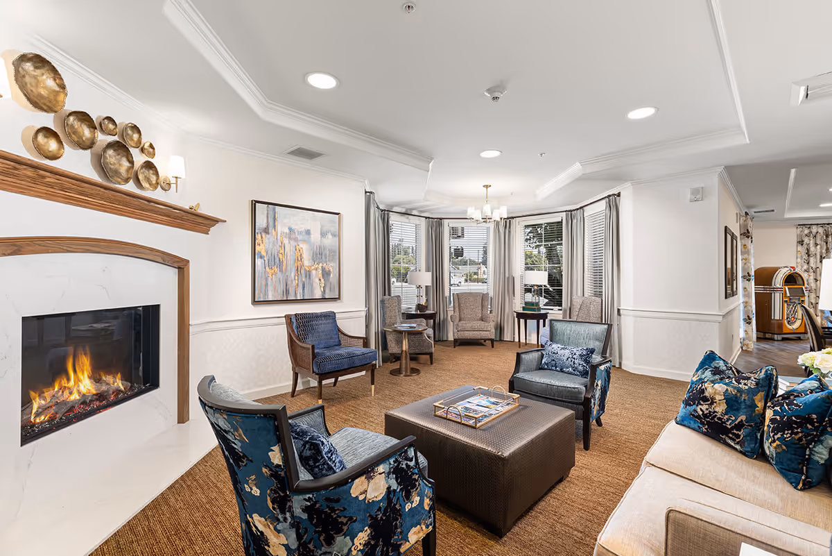 A cozy living room area with a lit fireplace on the left, surrounded by a white marble and wooden mantel. The room features a variety of seating including a beige sofa with blue patterned pillows, two blue floral armchairs, a dark ottoman coffee table with magazines, and additional chairs near the windows. The walls are white with crown molding, and there is a large abstract painting above the fireplace. The room is well-lit with recessed ceiling lights and a chandelier near the windows, which have white blinds and light curtains.