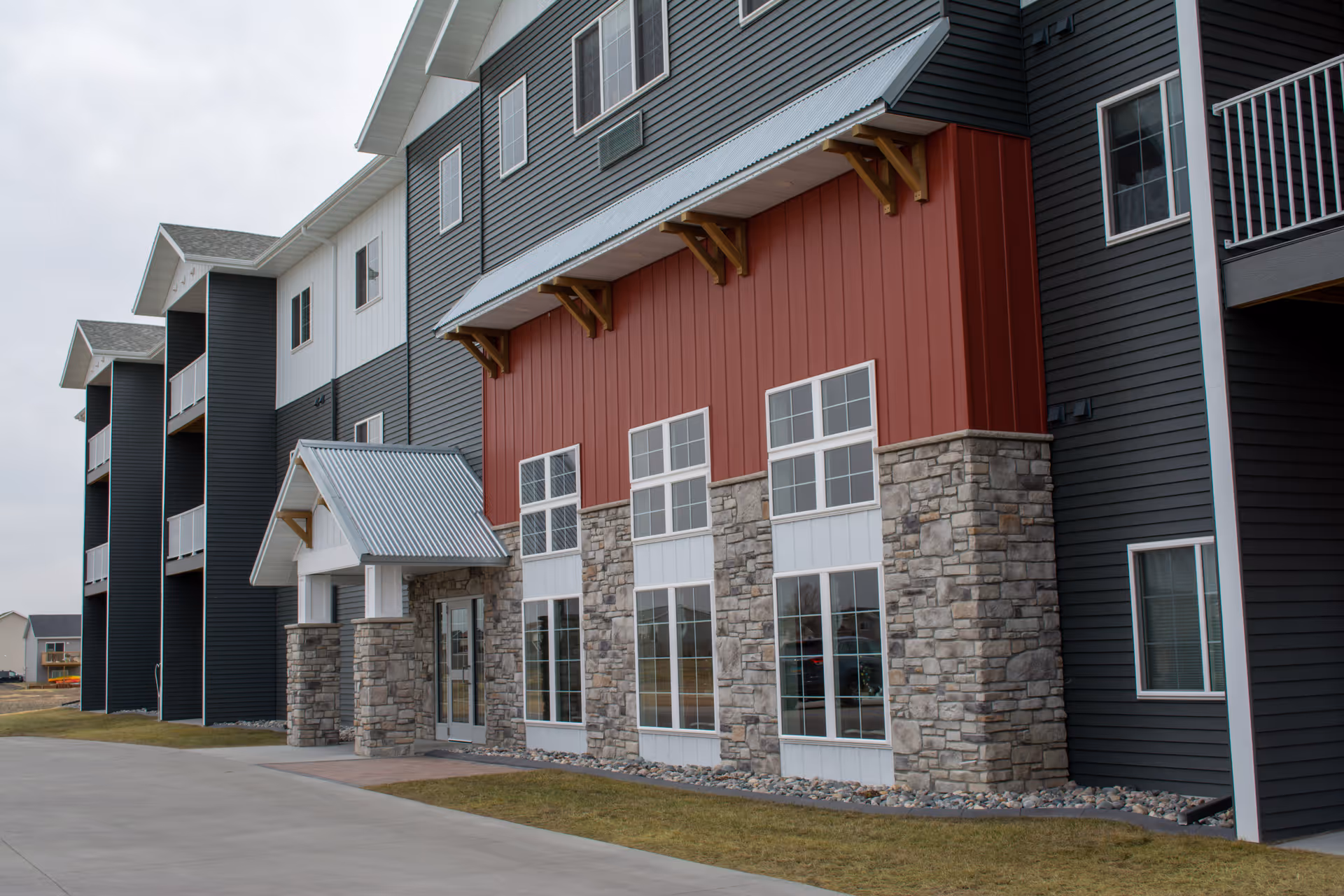 Exterior view of a modern multi-story building with a combination of dark siding, red paneling, and stone accents. The building has multiple windows and balconies, with a small covered entrance. The surrounding area includes a concrete driveway and a grassy lawn under an overcast sky.