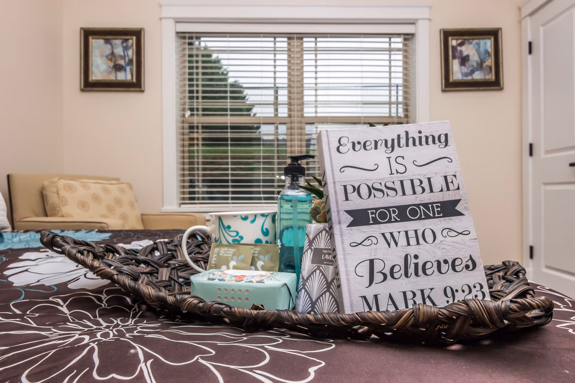 Woven tray holding a decorative sign, soap and toiletries on a patterned bedspread in a bedroom with a window and framed art.