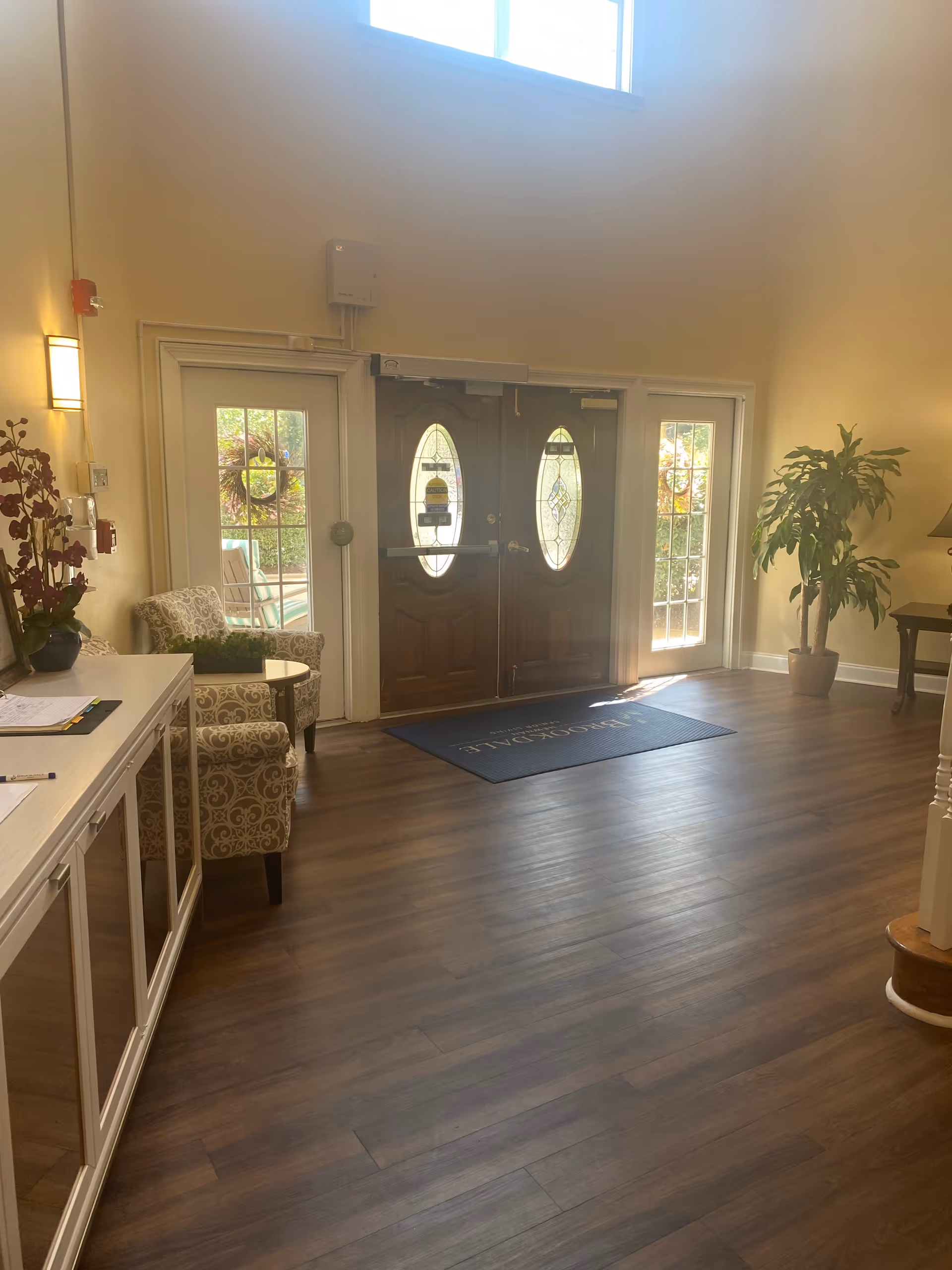 Interior view of a senior living facility entrance area with double wooden doors featuring oval glass windows. There are two patterned armchairs with a small round table between them on the left side, a white cabinet with papers and a pen on top, a potted plant on the right side, and a floor mat in front of the doors. The room has wooden flooring and cream-colored walls with a high window letting in natural light.