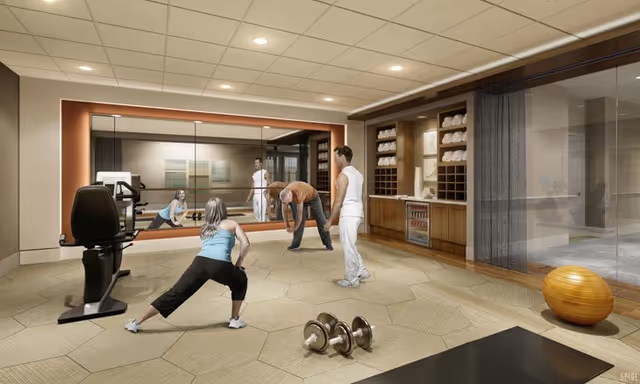 A fitness room with several people stretching and exercising in front of a large mirror, with gym equipment, weights, and towel storage visible.