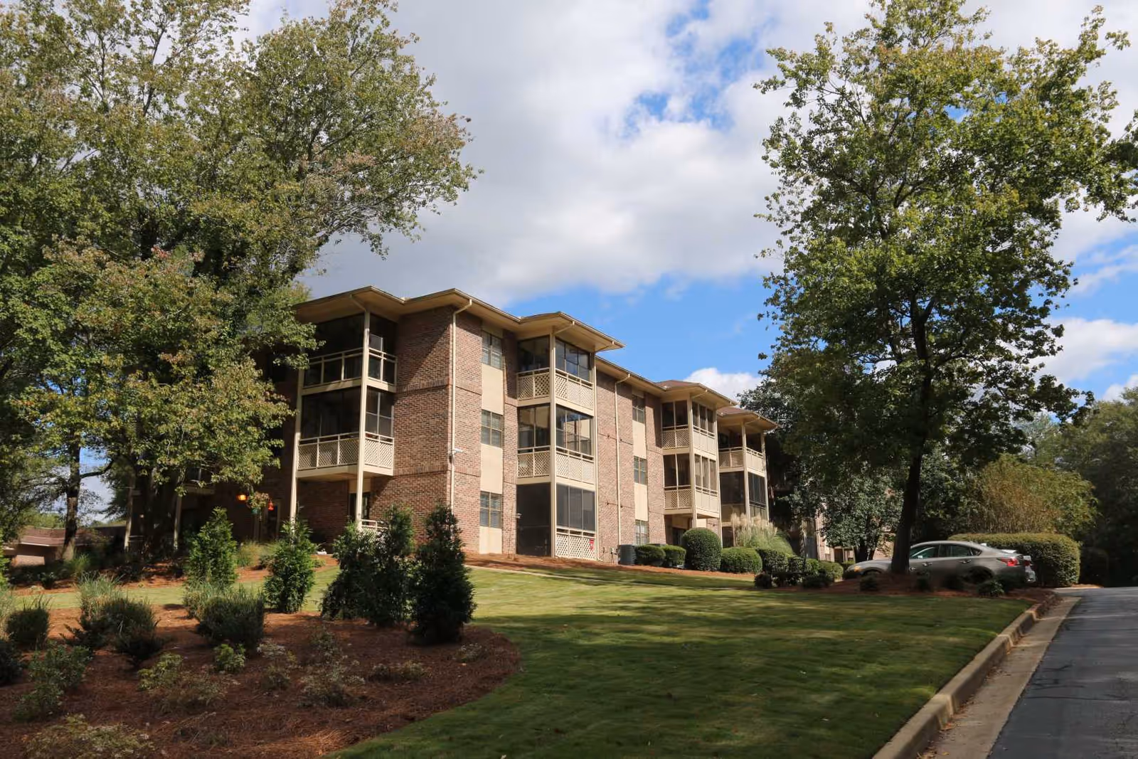 Three-story brick senior living building with screened balconies surrounded by trees and a landscaped lawn under a partly cloudy sky.