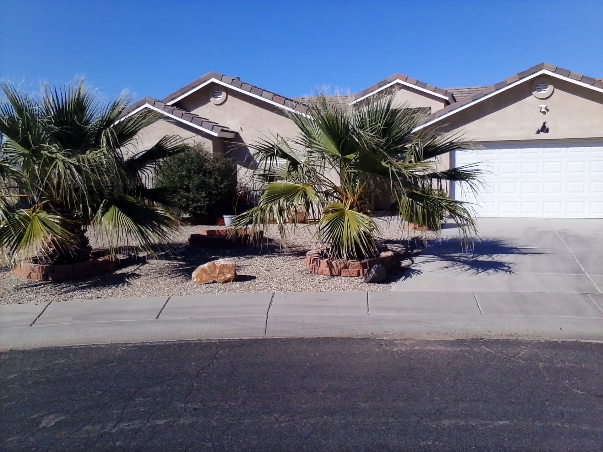 Front exterior view of a single-story house with a tiled roof, a white garage door, and desert landscaping featuring palm trees, bushes, rocks, and gravel under a clear blue sky.