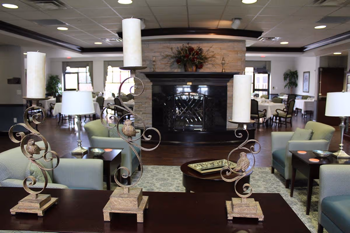 A cozy common area in a senior living facility featuring three decorative candle holders with bird designs on a wooden table in the foreground. Behind them are comfortable armchairs, side tables with lamps, and a large stone fireplace with a floral arrangement on the mantel. In the background, there are dining tables and chairs near windows letting in natural light.