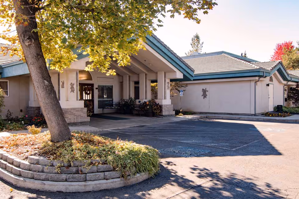 Exterior view of Edgewood Plantation Place showing the entrance with a covered driveway, a tree with green leaves in the foreground, and a small landscaped area with plants and flowers. The building has a light-colored facade with a teal roof trim and decorative wall art near the entrance.