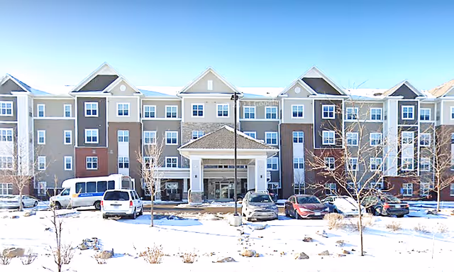 Front exterior view of a multi-story senior living facility building with a covered entrance, several parked cars, and leafless trees in a snowy landscape under a clear blue sky.