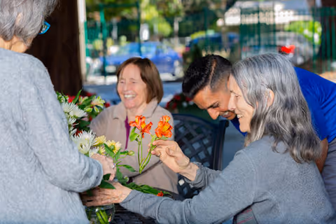 A group of elderly people and a caregiver are gathered around a table outdoors, arranging colorful flowers and smiling, enjoying a social activity in a garden or patio setting.