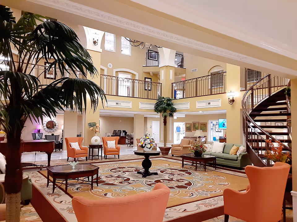 Spacious and elegant senior living facility common area with high ceilings, large windows, and a mezzanine level. The room is furnished with comfortable armchairs, sofas, coffee tables, and decorated with plants and floral arrangements. A spiral staircase is visible on the right side, and a grand piano is on the left.