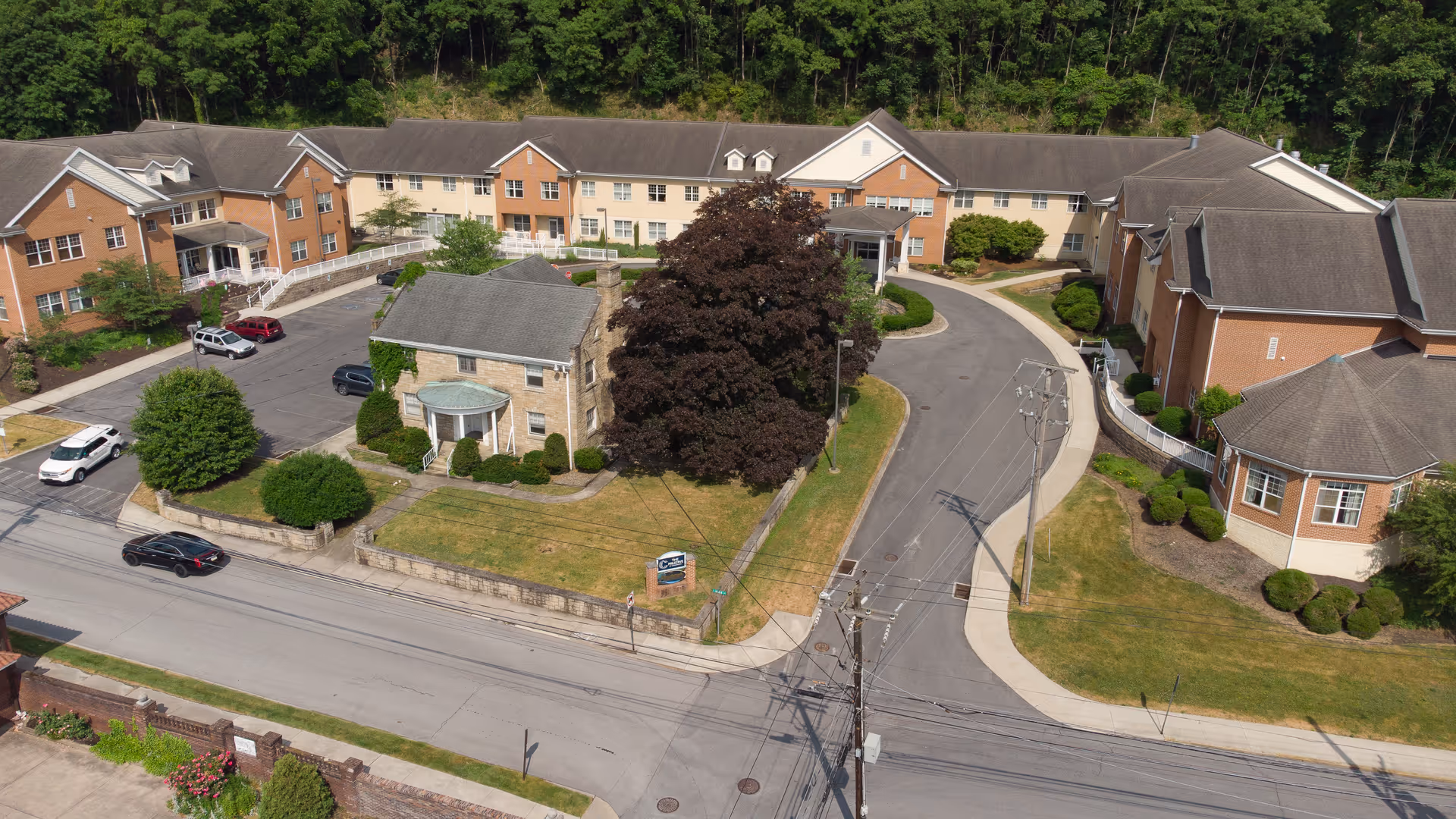 Aerial view of a U-shaped brick and beige multi-story senior care building with a curved driveway, parking area, lawns, and large trees.