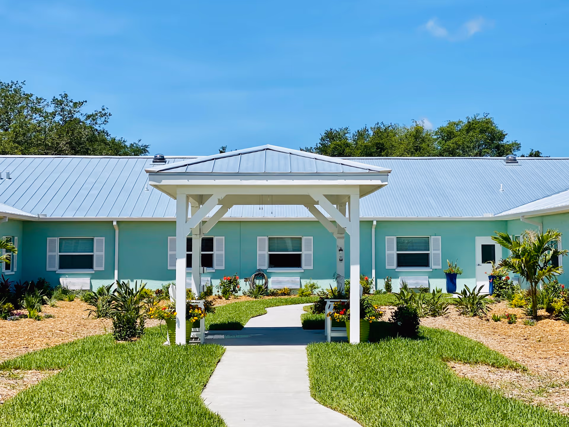 Outdoor view of Trinity Place Assisted Living showing a light blue building with a metal roof, white window shutters, and a white pergola over a concrete pathway surrounded by green grass and landscaped plants and flowers under a clear blue sky.