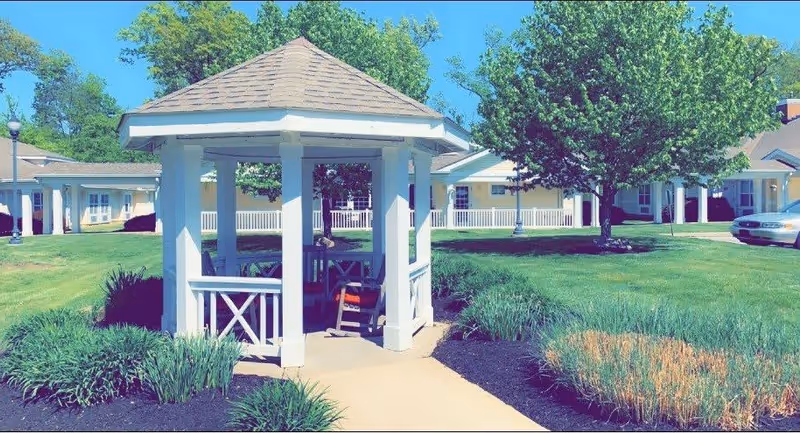 A white gazebo with a shingled roof sits on a paved walkway surrounded by green grass, bushes, and trees in an outdoor garden area of a senior living facility. In the background, there are single-story buildings with white railings and a parked car on the right side.