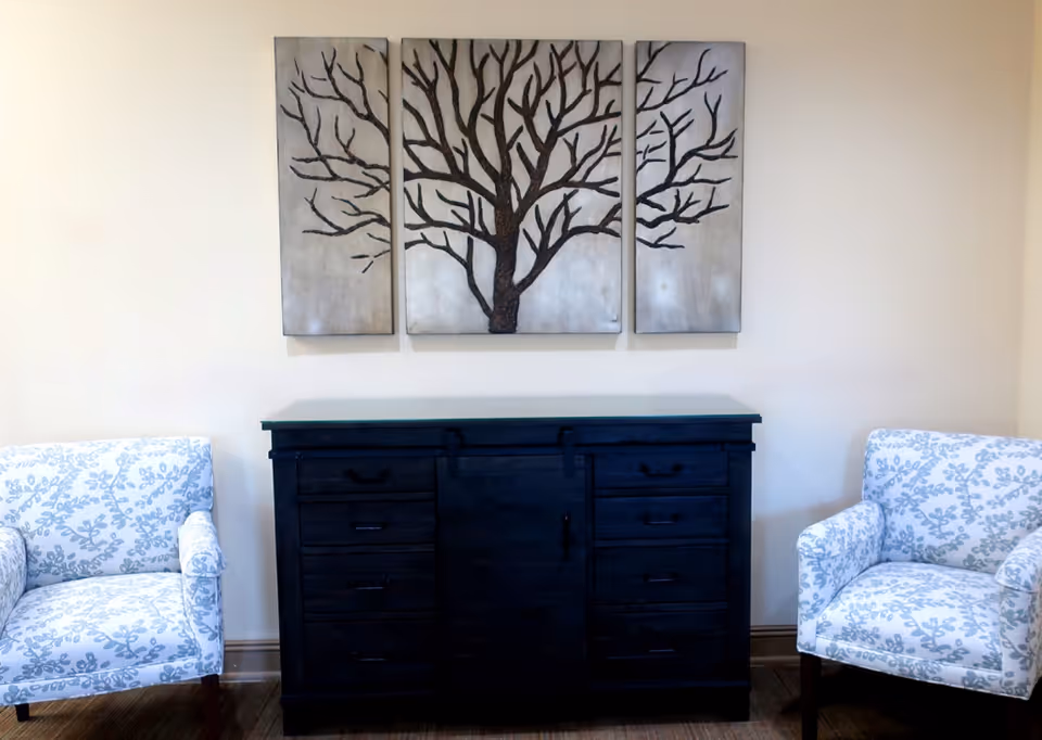 A living room area with two white armchairs featuring a blue floral pattern placed on either side of a dark wooden cabinet with multiple drawers and a sliding door. Above the cabinet, there is a three-panel wall art depicting a tree with bare branches on a light background.