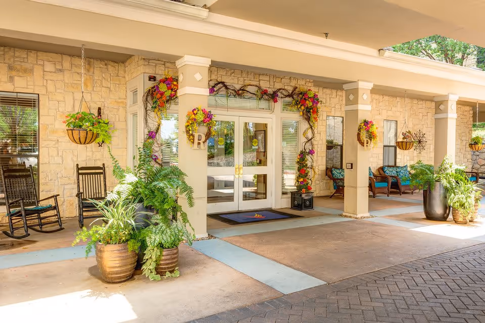 Entrance area of Ridgmar Place featuring a covered porch with stone walls, double glass doors decorated with floral wreaths, hanging flower baskets, potted plants, rocking chairs, and cushioned wicker seating.