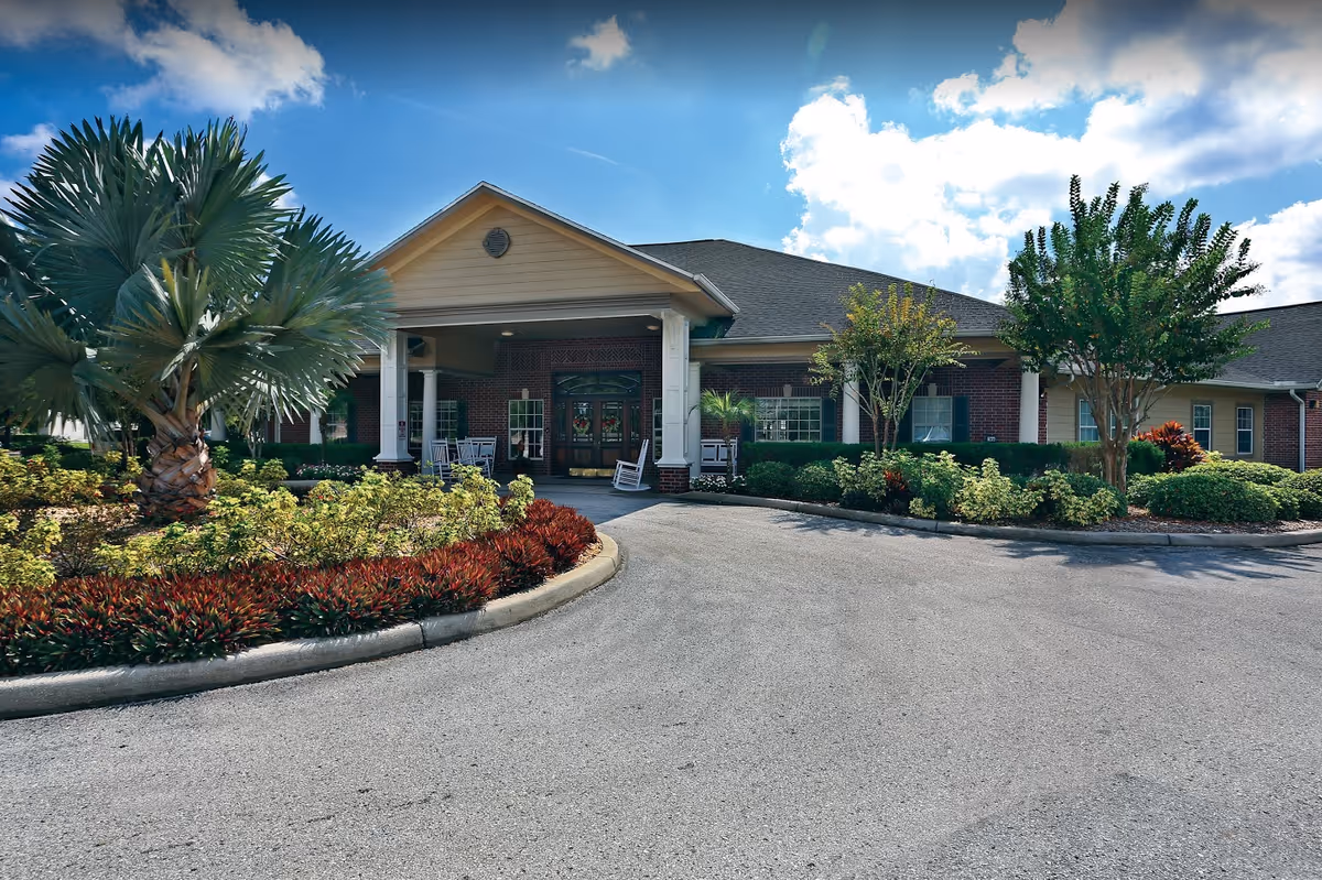 Exterior view of Sodalis Tampa facility showing a single-story building with a covered entrance supported by white columns. The driveway curves in front of the entrance, surrounded by landscaped greenery including palm trees, bushes, and other plants under a partly cloudy blue sky.