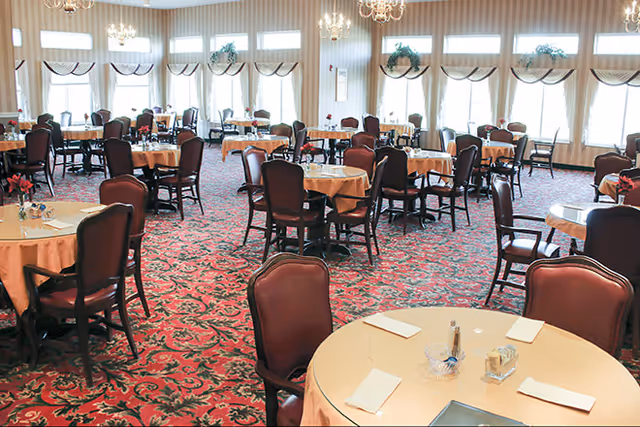 A spacious dining room with multiple round tables covered with beige tablecloths, each surrounded by dark wooden chairs with cushioned seats. The room features large windows with sheer curtains and valances, allowing natural light to fill the space. Elegant chandeliers hang from the ceiling, and the floor is covered with a red and green patterned carpet.