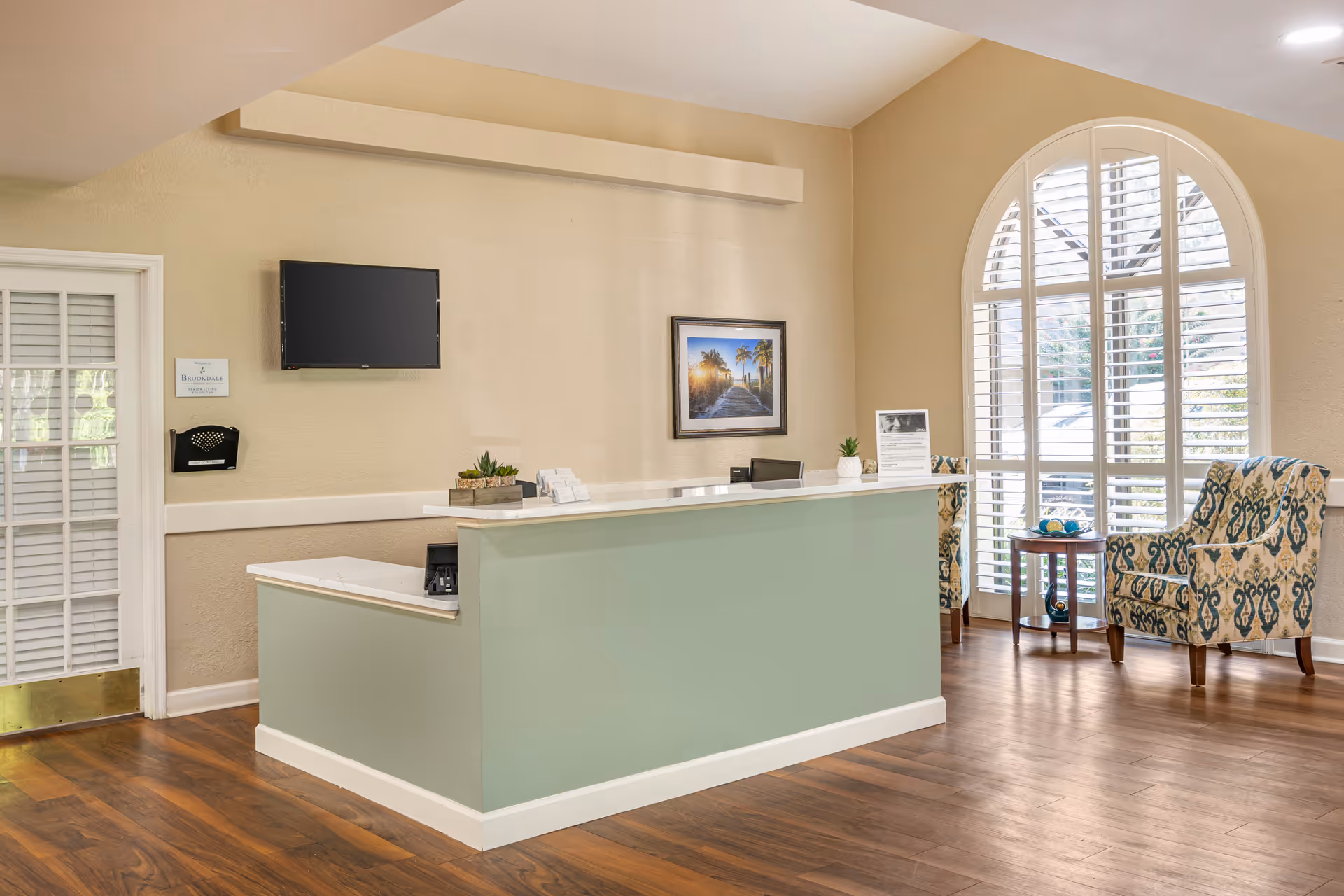 Bright reception lobby with a mint-green front desk, patterned armchairs, a large arched window with shutters, and a wall-mounted TV.