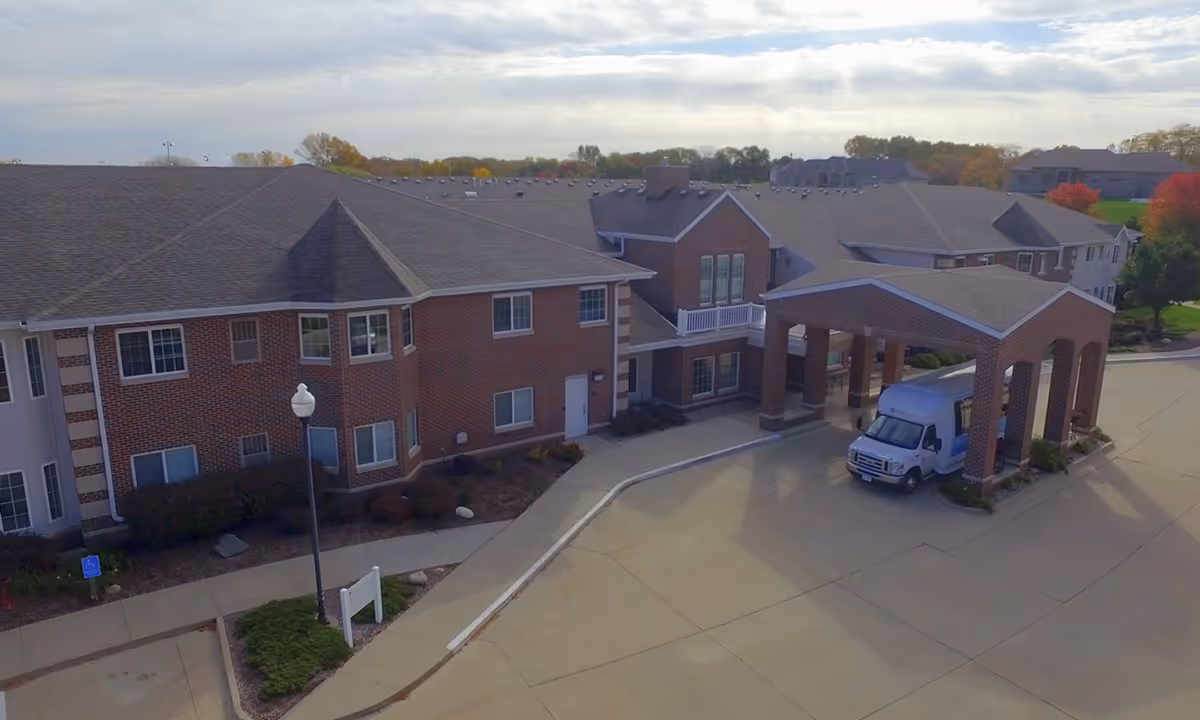 Front entrance of a two-story brick senior living building with a covered porte-cochère and a parked shuttle van.