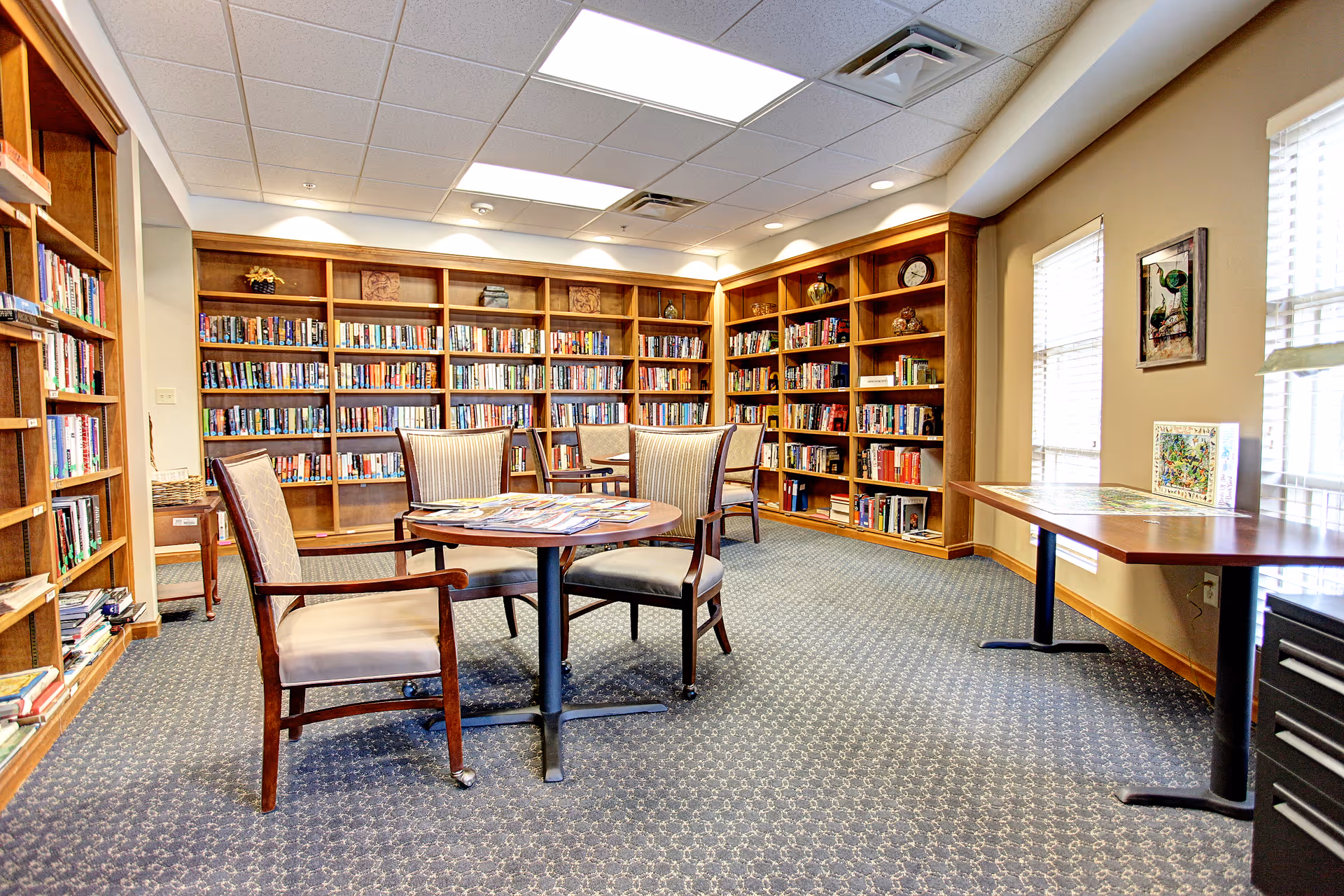 A cozy library room with wooden bookshelves filled with books lining the walls. There are several cushioned chairs around a round table in the center of the room. The room has carpeted flooring, a drop ceiling with fluorescent lights, and windows on the right side letting in natural light. A rectangular table with a puzzle on it is positioned near the windows.