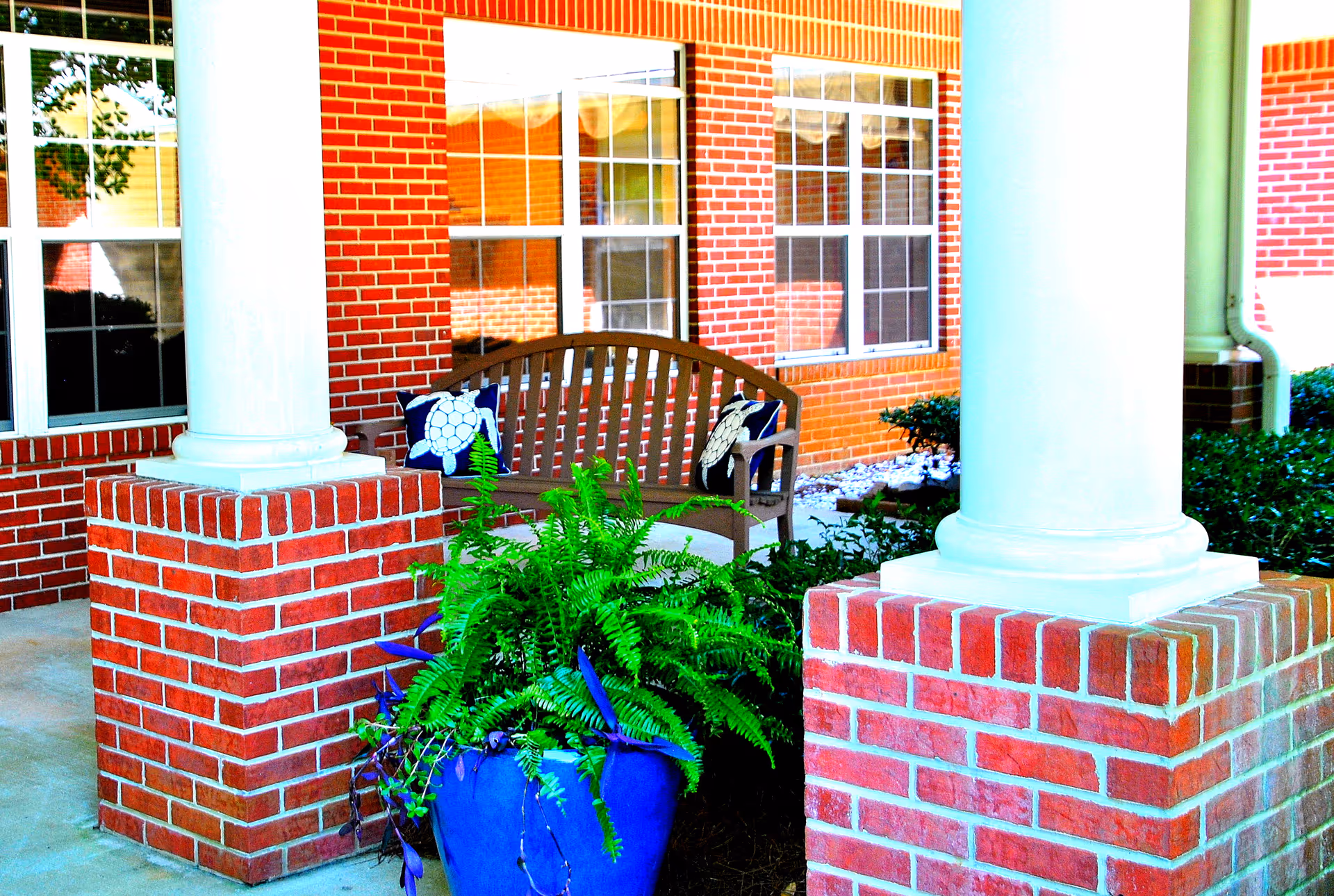 Front porch of a brick building with white columns, a bench, and a blue planter of ferns.