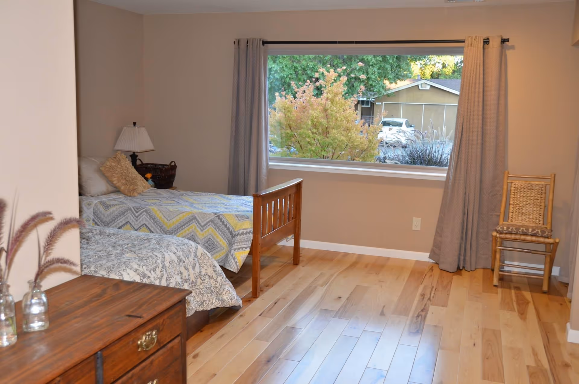 A bedroom with two single beds, one with a patterned yellow and gray bedspread and the other with a white and gray bedspread. There is a wooden nightstand with a lamp and a basket between the beds. A large window with gray curtains shows greenery and a house outside. A wooden chair is placed near the window, and a wooden dresser with decorative vases is in the foreground.
