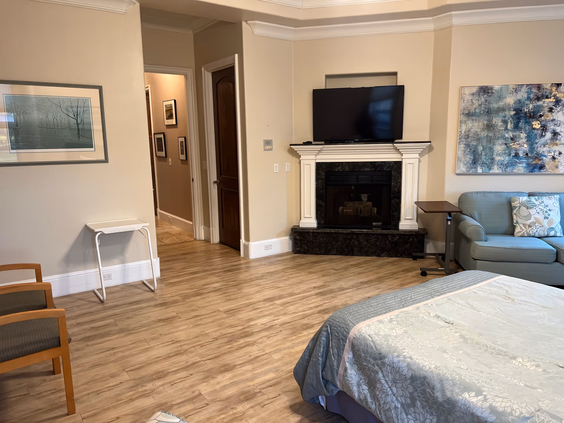 Spacious senior living suite with a bed in the foreground, a seating area with couch and chairs, and a fireplace with a TV above on wood-look flooring.