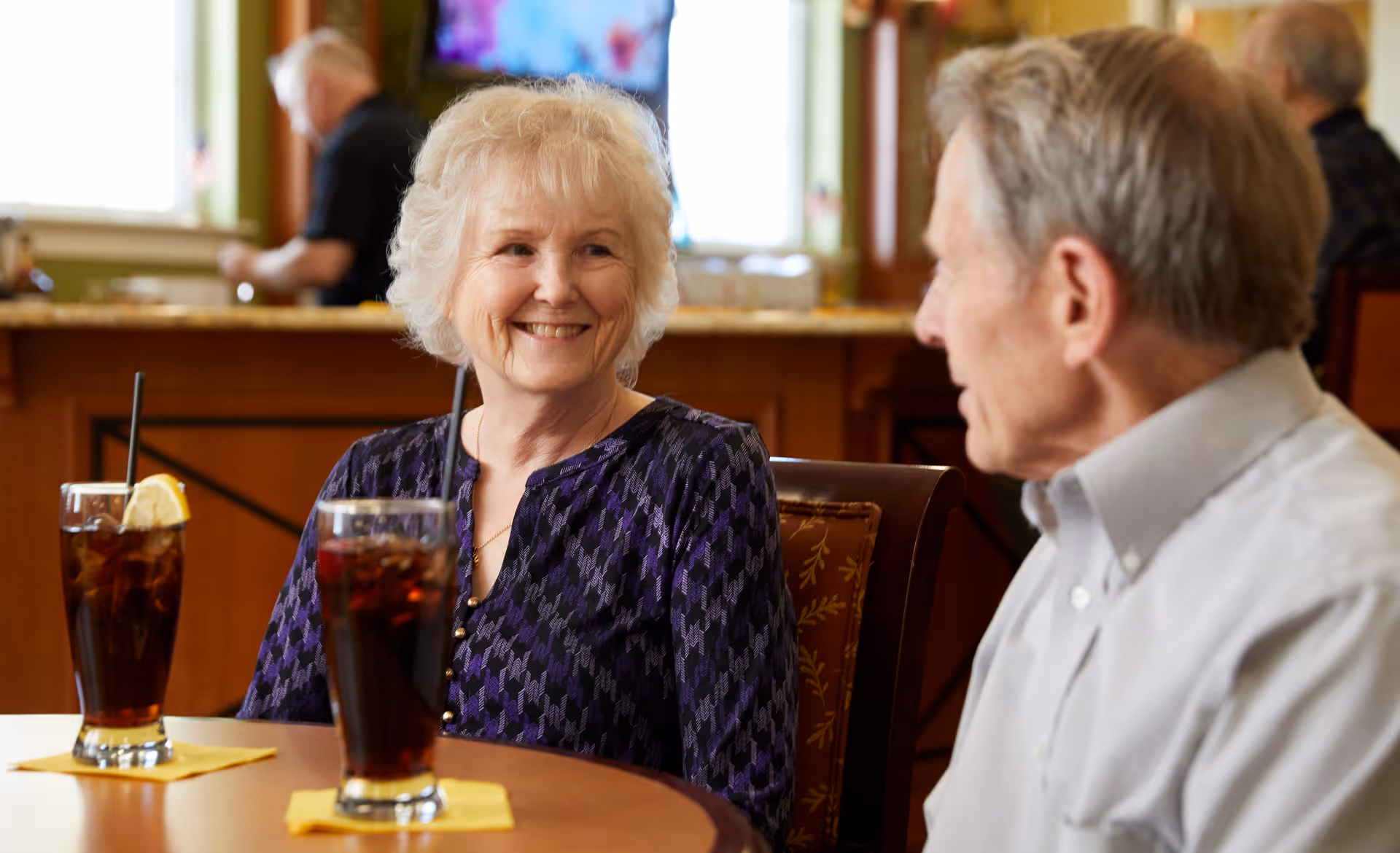 An elderly woman and man sitting at a table in a dining area, smiling and talking. Two glasses of iced tea with lemon wedges and straws are on the table in front of them. The background shows a counter and another person standing near a window.