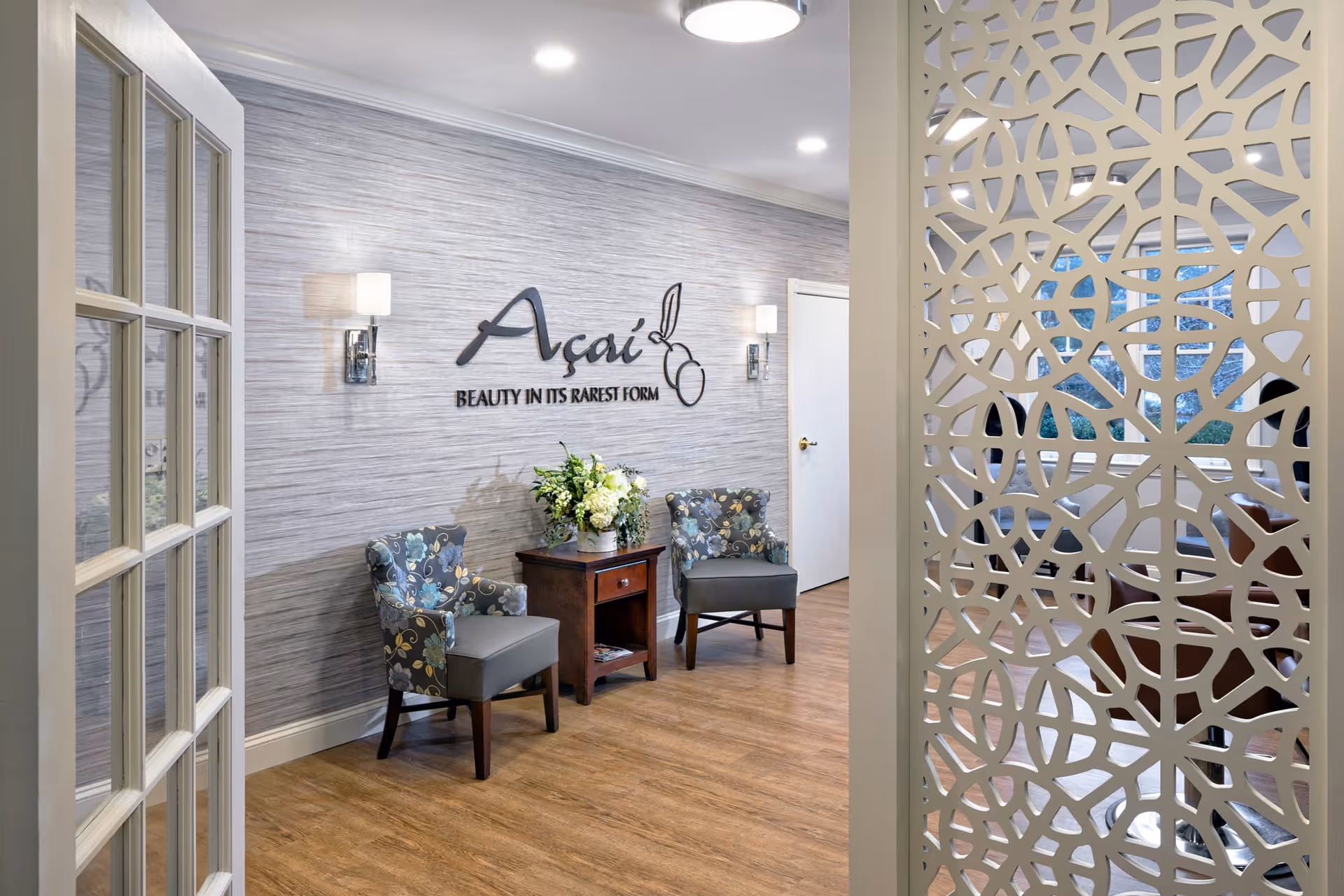 Interior view of a waiting or lounge area with two patterned armchairs and a wooden side table with a flower arrangement. The wall behind has a sign that reads 'Açaí BEAUTY IN ITS RAREST FORM'. The space has wood flooring, modern wall sconces, and a decorative white partition partially visible on the right side.
