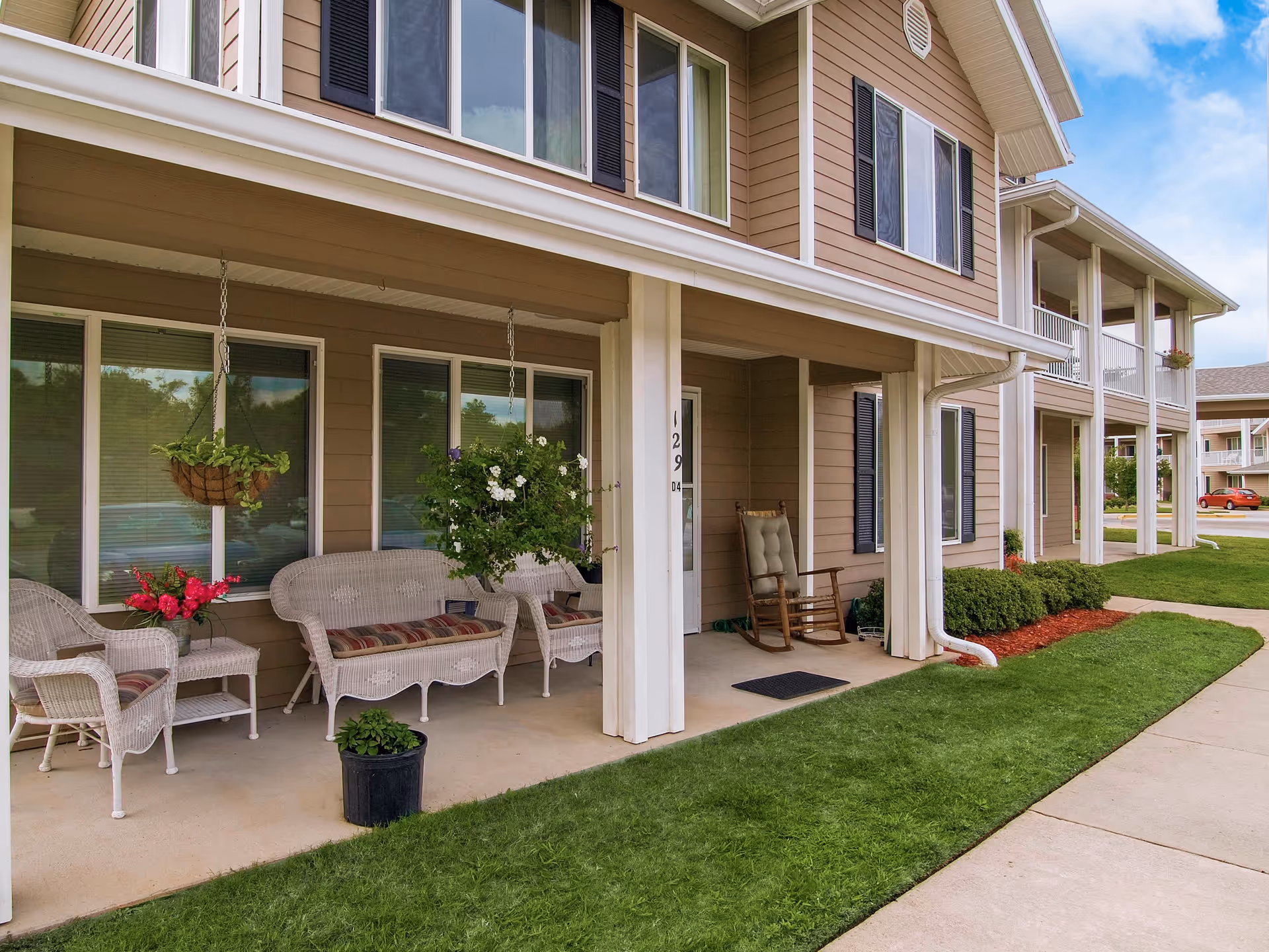 Covered porch area of a senior living facility with white wicker chairs and a loveseat with cushions, a rocking chair, hanging flower baskets, potted plants, and windows with black shutters. The building exterior is beige with white trim, and there is a sidewalk and green lawn in front.