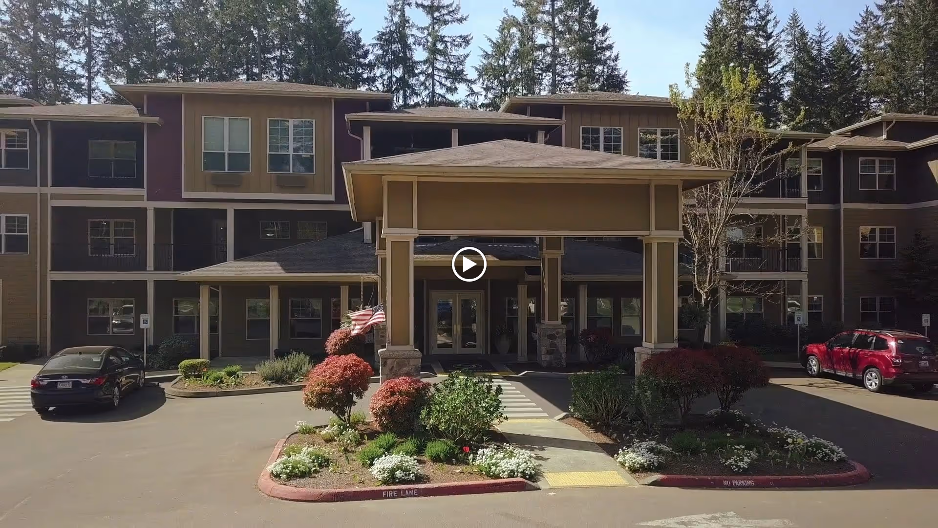Front exterior view of a multi-story senior living facility with a covered entrance, landscaped garden beds with shrubs and flowers, an American flag, and parked cars on either side of the driveway.