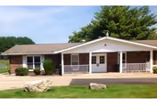 Single-story facility exterior with brick and white siding, porch entrance, and green lawn.