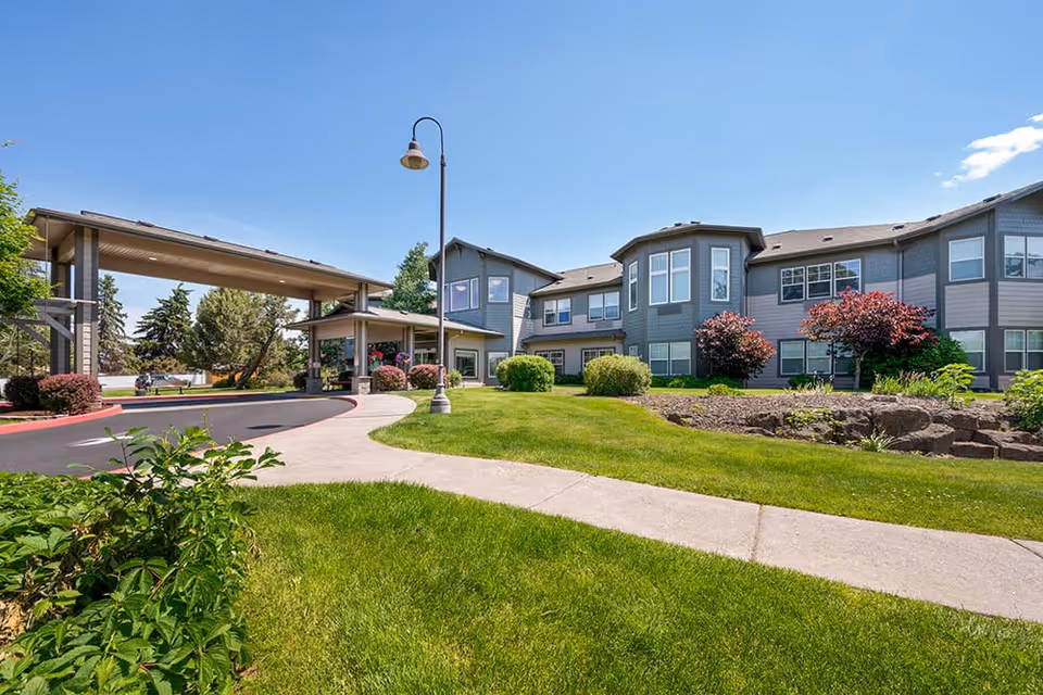 Exterior view of Brookdale Redmond senior living facility on a sunny day, showing a two-story building with gray siding, multiple windows, landscaped green lawn, bushes, and a covered driveway entrance with a street lamp.