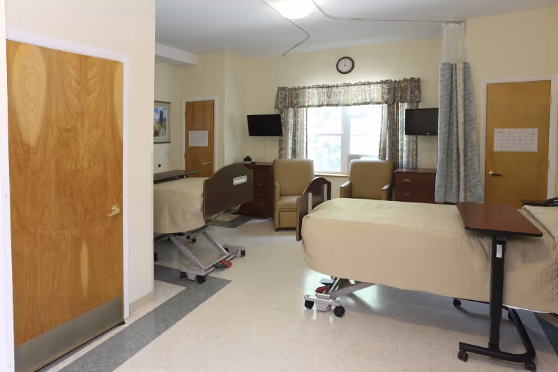 A nursing facility room with two hospital beds, two beige armchairs, two small wooden dressers, two wall-mounted televisions, a window with floral curtains, a clock above the window, and two wooden doors. The room has a light-colored floor and walls.