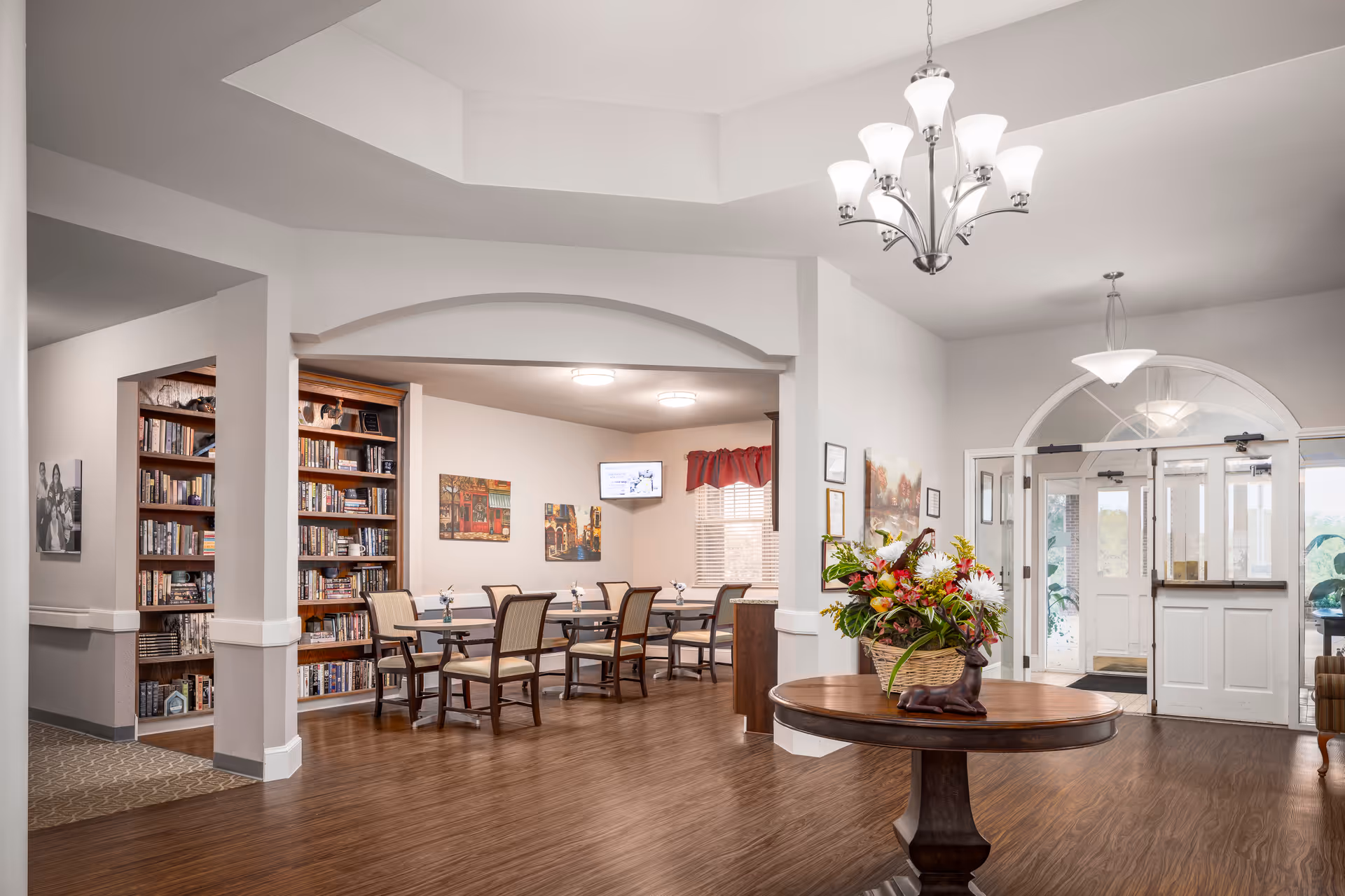 Interior view of a senior living facility showing a common area with a round wooden table holding a flower arrangement in the foreground. In the background, there is a dining area with tables and chairs, bookshelves filled with books, wall art, and a wall-mounted TV. The entrance doors with glass panels are visible on the right side.