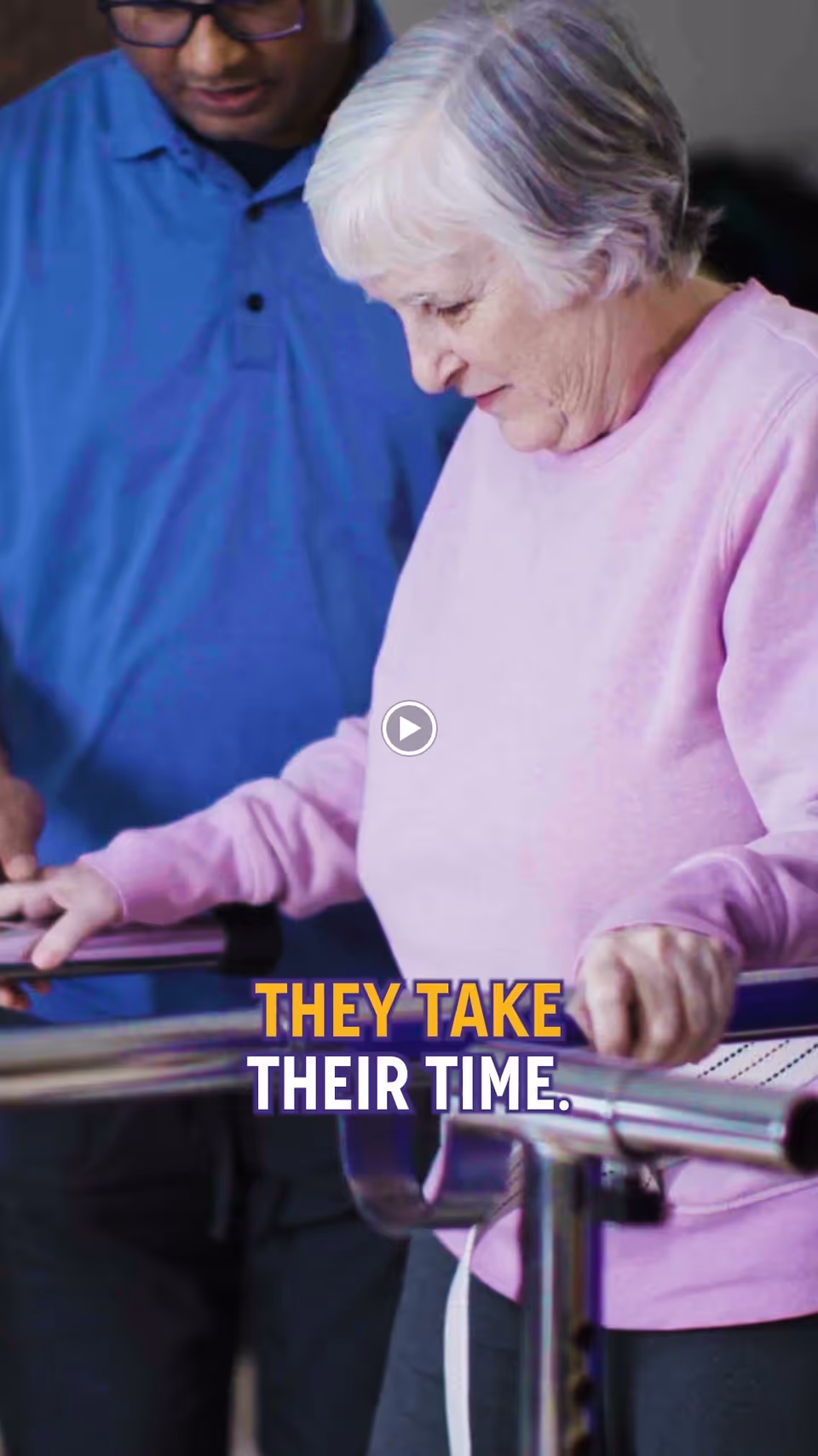 An elderly woman in a pink sweater uses parallel bars for walking assistance while a caregiver in a blue shirt stands nearby, providing support and encouragement.