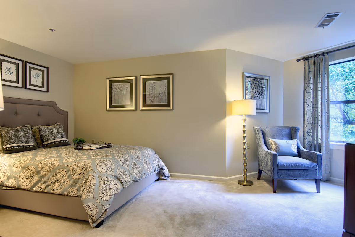 Well-lit bedroom with a patterned bed, decorative pillows, framed artwork, an upholstered armchair by a window, and a floor lamp.