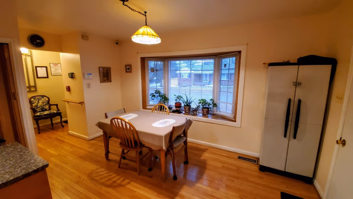 Dining room with a table covered by a cloth and four chairs under a pendant light, a bay window with potted plants, and a tall storage cabinet.