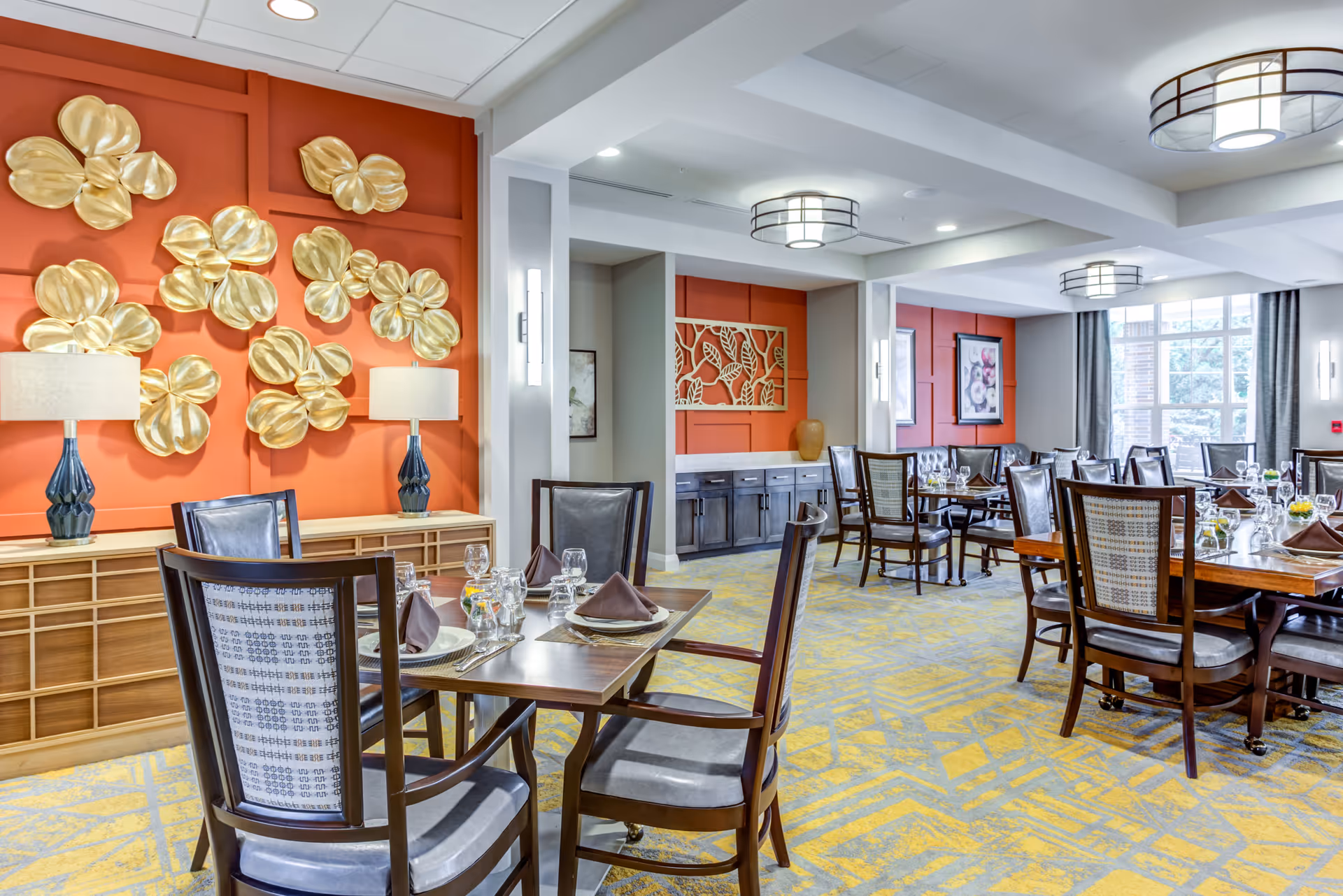 A bright and elegant dining room with wooden tables and chairs set with plates, glasses, and brown napkins. The walls are painted orange with decorative gold floral wall art and framed pictures. The room has modern ceiling lights and large windows with gray curtains letting in natural light. The carpet has a yellow and gray geometric pattern.