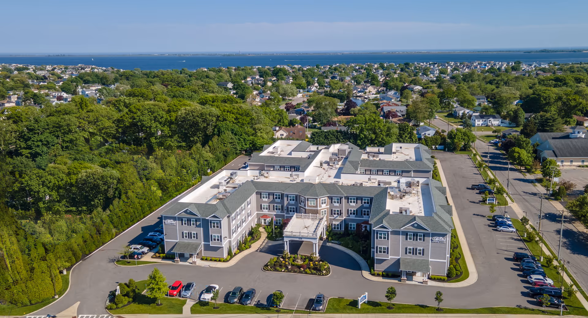 Aerial view of a three-story assisted living building with a circular front drive, parking lots, surrounding trees, and a distant coastline.