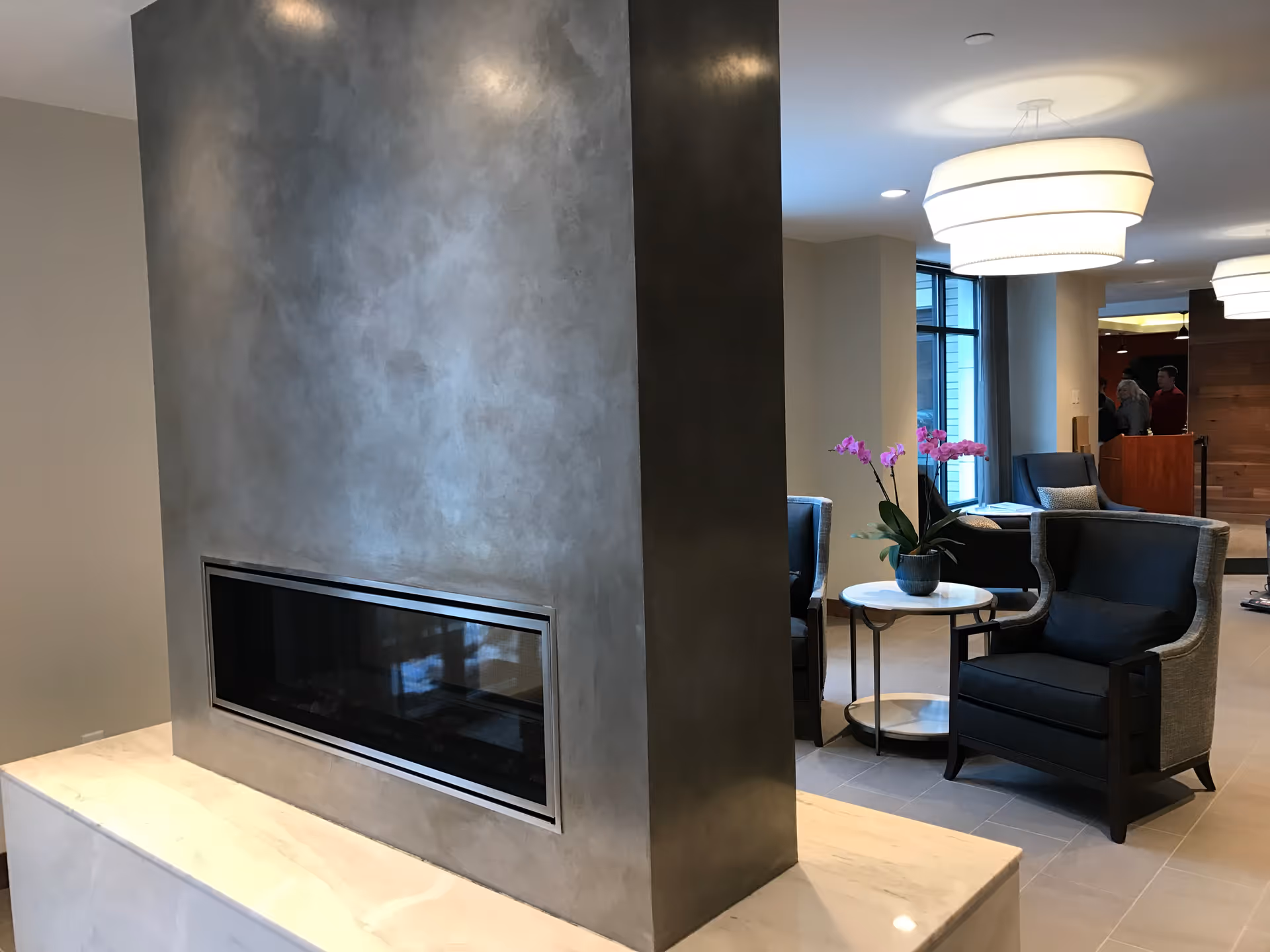 Modern communal lounge with a built-in horizontal fireplace, armchairs, a side table with an orchid, and pendant lights.