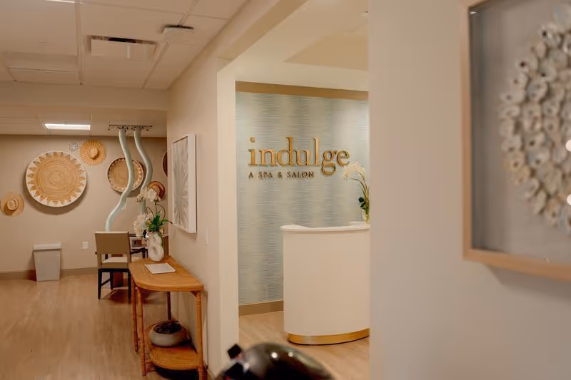 Interior view of a spa and salon reception area with a sign reading 'indulge A SPA & SALON' on a textured blue wall. The space features light wood flooring, a white curved reception desk, decorative wall art, and a small table with a vase and flowers in the foreground.