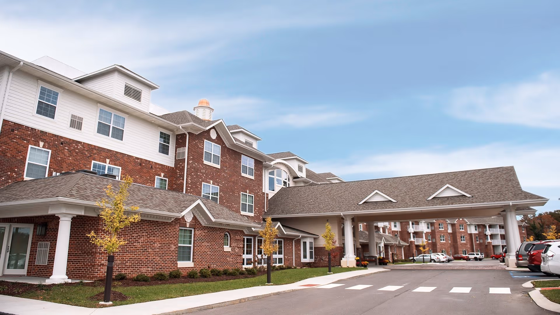 Exterior view of a multi-story brick and white siding building with a covered entrance driveway, small trees, and parked cars under a partly cloudy sky at Ashwood Square Retirement Community.