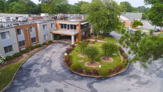Aerial view of the Millennium Post Acute Rehabilitation facility showing a curved driveway around a landscaped circular island with small trees and shrubs, surrounded by a multi-story building and parking area with cars and greenery in the background.