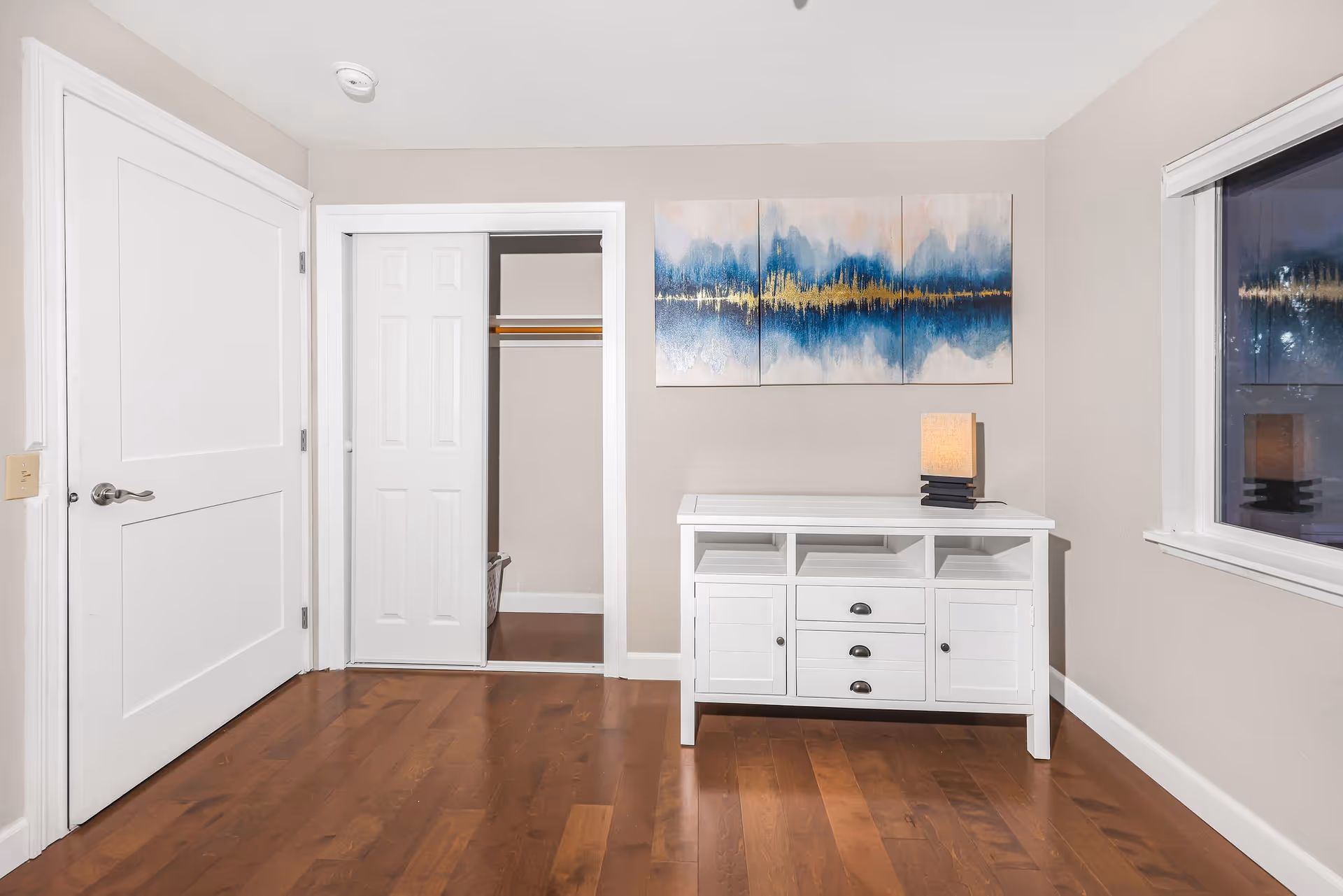 A room with wooden flooring featuring a white door on the left, a closet with sliding white doors in the center, and a white cabinet with drawers and shelves on the right. Above the cabinet is a three-panel abstract painting in blue, gold, and beige tones. A small lamp with a beige shade sits on the cabinet. There is a window on the far right wall.