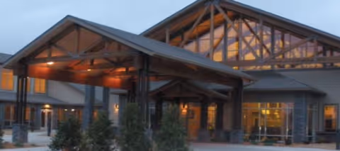 Exterior view of RiverLodge Assisted Living facility at dusk, showing a large covered entrance with wooden beams and glass windows illuminated from inside.