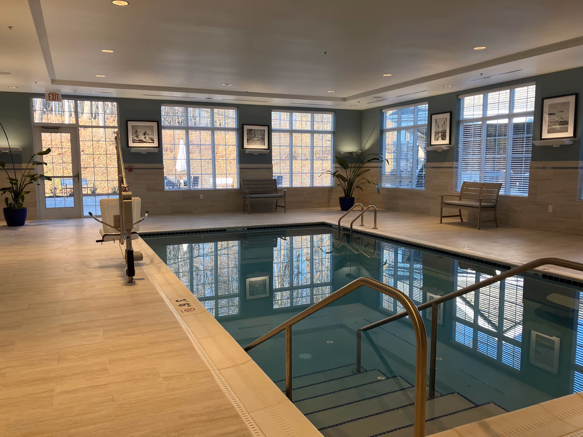 Indoor swimming pool area with large windows letting in natural light, beige tiled floor and walls, two benches, potted plants, and a pool lift for accessibility.