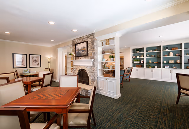 A spacious senior living common area featuring a stone fireplace with a painting above it, several wooden tables with chairs, built-in white shelves filled with decorative items, and soft lighting creating a warm and inviting atmosphere.