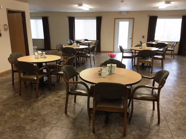 A dining room with several round tables and chairs arranged neatly. Each table has a small centerpiece and condiments. The room has large windows with dark curtains and a door leading outside. The floor is tiled and the walls are light-colored with wooden trim.