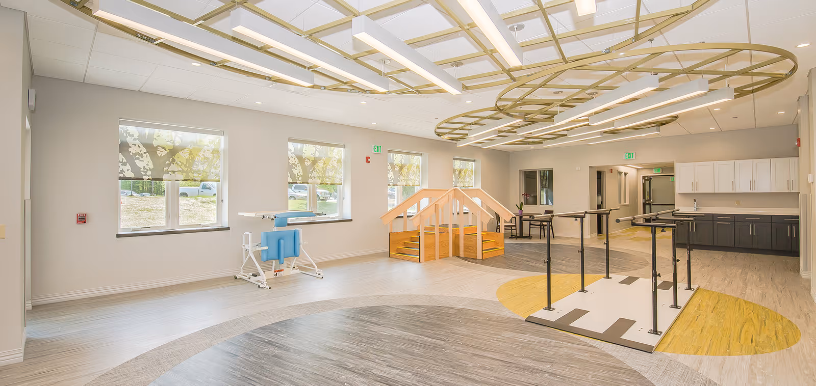 A spacious rehabilitation or therapy room with large windows covered by patterned blinds, a small wooden staircase with handrails, parallel bars for walking exercises, a blue therapy chair, and cabinetry along the back wall. The ceiling has modern circular light fixtures and the floor has a mix of wood and vinyl patterns.