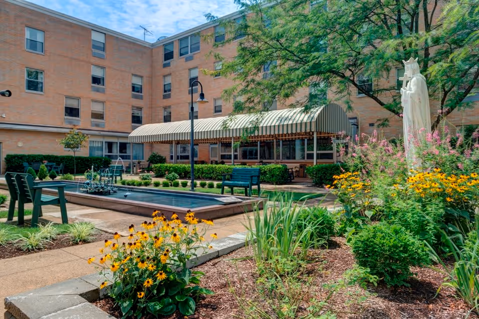 Outdoor courtyard area of a senior living facility with a rectangular water fountain, green benches, a variety of flowering plants, and a white statue of a crowned figure holding a cross. The building surrounding the courtyard is made of brick with multiple windows, and there is a covered patio area with striped awnings.