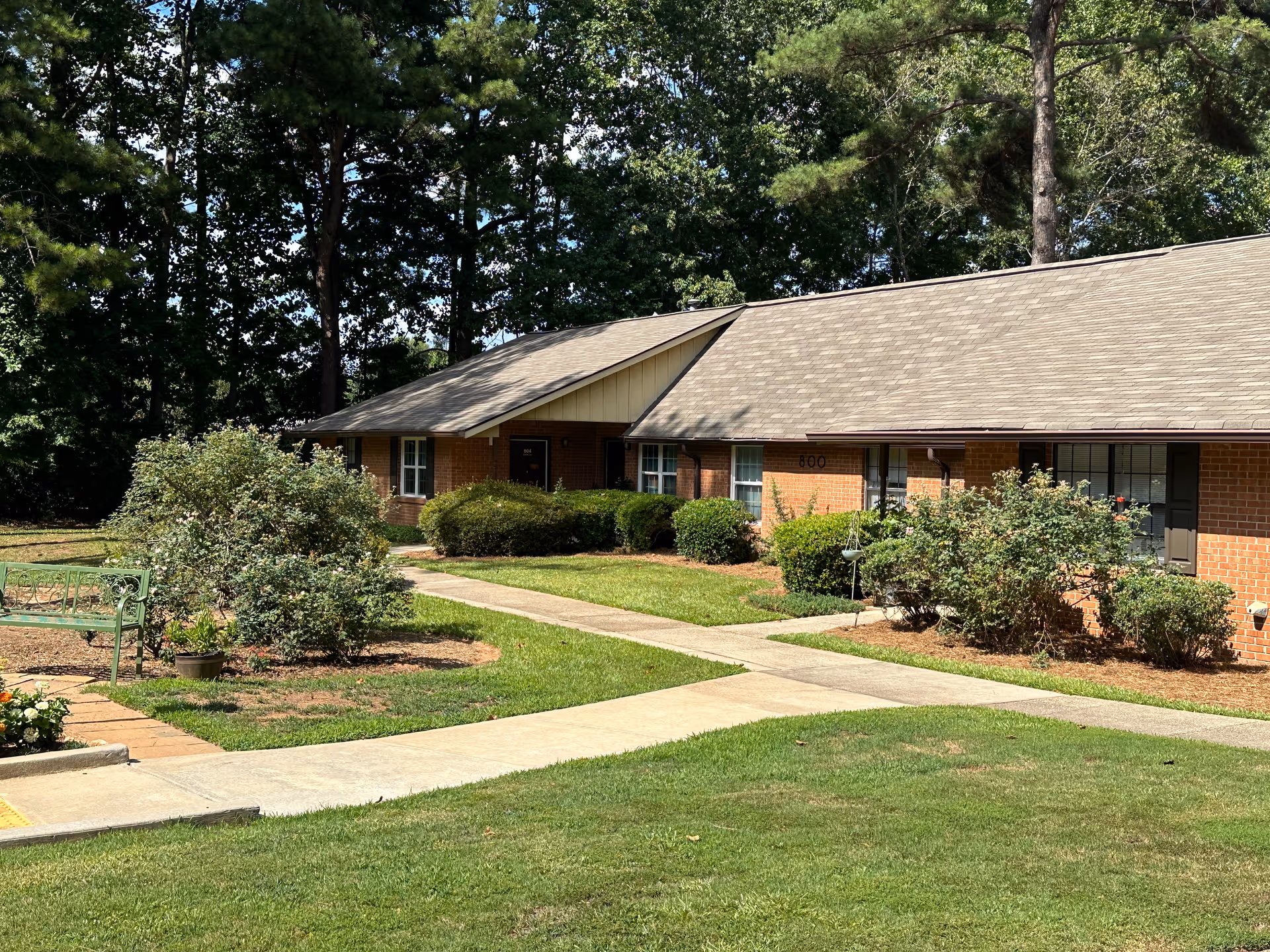Single-story brick residential building with a sloped roof surrounded by green grass, bushes, and trees. A concrete walkway leads to the entrances of the units. There is a green metal bench and some potted plants near the walkway.